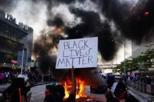 Mężczyzna trzymający tablicę z napisem Black Lives Matter obok samochodu policyjnego płonącego w czasie protestu przed budynkiem CNN Center 29.05.2020 r. w Atlancie w stanie Georgia<br/>(Elijah Nouvelage / Getty Images)
