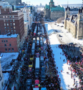 Ciężarówki tworzące Freedom Convoy 2022 stoją na ulicy, podczas gdy ludzie niosą tablice z hasłami przeciwko rządowi, Ottawa przed Parliament Hill, Kanada, 29.01.2022 r. (ANDRE PICHETTE/PAP/EPA)