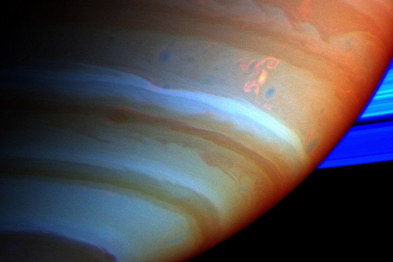Smocza burza. Zdjęcie wykonane 15.09.2004 r. przez sondę Cassini. Atmosfera Saturna i jego pierścienie pokazane są tutaj przy użyciu sztucznie dobranych kolorów, kompozycja wykonana ze zdjęć Cassini zrobionych w bliskiej podczerwieni przez filtry wykrywające różne ilości metanu. Części atmosfery z dużą ilością metanu ponad chmurami są czerwone, co wskazuje na chmury znajdujące się głęboko w atmosferze. Szary kolor oznacza wysokie chmury, a brązowy oznacza chmury na średnich wysokościach (NASA/JPL/Space Science Institute / zdjęcie modyfikowane, <a href="https://commons.wikimedia.org/w/index.php?curid=1181461">domena publiczna</a>)